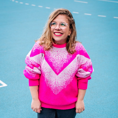 Ninia in einem pinken Pullover steht auf einem blauen Sportfeld und schaut lächelnd in den Himmel